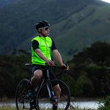A person cycling on a road wearing a high visibility yellow cycling vest with reflective piping, in front of a mountainous landscape.