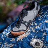 A pair of UDOG Distanza gravel bicycle shoes featuring a carbon cinder black and ash grey color scheme, positioned on a rocky surface with a blue background.