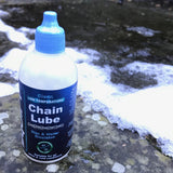 A bottle of Clean Chain Lube low-temperature chain lube with a blue cap, placed on a wet concrete surface with patches of snow.