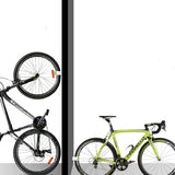 Two mountain bikes hanging on a wall-mounted bicycle rack, which is in a vertical position. The rack is mounted on a black pole, and one bike is positioned above the other with a space gap between them.