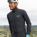 A person wearing a black Tineli Pro Rain Jacket, with reflective details visible on the sleeves. The jacket has a slim fit and is designed for cycling, showing no hood and featuring adjustable cuffs.